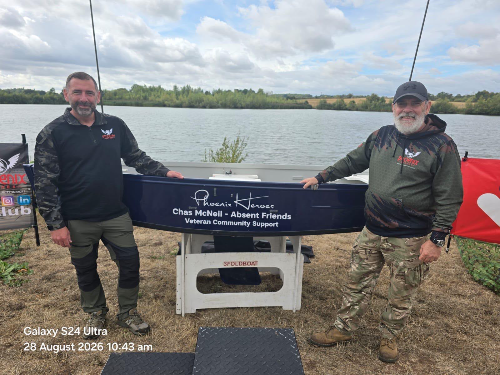 Two Phoenix Heroes veterans at the 2026 Veterans Raffle Shield beside a boat lettered Chas McNeil, Absent Friends